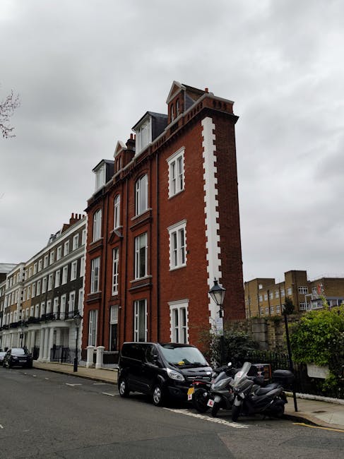 A multi-storey red brick building with white window frames and decorative white accents on a city street. The building has six visible floors, including a small attic level with dormer windows, and features a pitched roof. On the street in front, there are black cars and parked scooters close to the curb. The street is lined with similar Victorian-style terraced houses, and a lamppost is situated near the building. The sky above is overcast with gray clouds, creating a subdued lighting environment typical of a typical urban residential area. This scene illustrates an inner-city neighbourhood, potentially relevant for house removals and moving services, highlighting the type of properties often handled by companies like Man with Van West Kensington.