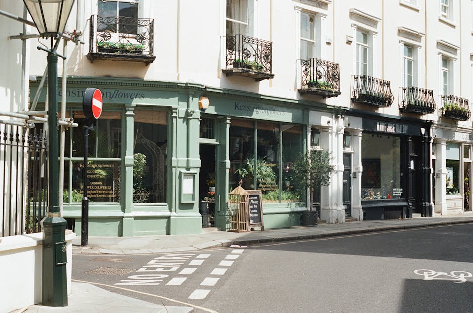 The image shows a street scene outside a building featuring a green, traditional-style shopfront with large glass windows displaying plants and flowers. The shop, with the sign 'Kensington Flowers', is situated on the corner of a pavement, with a corner post and a no-entry road sign nearby. Adjacent to the shop, to the right, is a cream-colored building with multiple floors, each with small balconies with decorative wrought iron railings and potted plants. The sidewalk includes a small wooden easel next to the shop entrance, and the street is paved with markings indicating a bicycle lane. A metal railing separates the pavement from the road, which is free of traffic. The scene is lit with natural daylight, suggesting a clear or partly cloudy day, capturing a typical London street view relevant to local property and business premises, often associated with house and commercial removals or relocation services provided by companies such as Man with Van West Kensington.