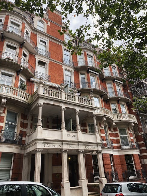 A multi-storey residential building with a classical architectural style, featuring red brick walls and ornate white stone balconies with decorative balustrades. The building has several windows, some with white window frames and others with black wrought iron railings, and is partially obscured by green leafy trees in the foreground. At the ground level, there is a white stone entrance with several columns supporting a balcony above, inscribed with the address '13-36 CAMPDEN HILL COURT.' Parked vehicles are visible at the base of the building on the street. This scene illustrates a typical property where house removals and furniture transport could take place, fitting the context of house moving services offered by Man with Van West Kensington during packing, loading, and home relocation processes.
