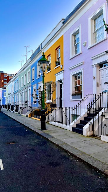 A row of colorful terraced houses on Fulham Palace Road in West Kensington, W14, with facades painted in pastel shades of pink, yellow, blue, and purple. The houses feature white-framed sash windows, small front gardens enclosed by black metal railings, and concrete steps leading to front doors. A vintage-style street lamp is positioned on the pavement in front of one of the houses. The sky is clear with natural daylight illuminating the scene, highlighting the neat and well-maintained appearance of the properties. This residential street is typical of West Kensington, with a mix of old and modern styles, and no visible signs of activity or moving equipment present in the image. The scene provides a calm setting suitable for home relocation or furniture transport services conducted by companies such as Man with Van West Kensington.