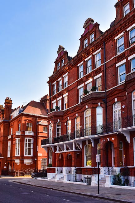 A multi-storey red-brick Victorian building with white decorative accents, large sash windows, and black wrought iron balconies on the upper floors, situated on Fulham Palace Road in West Kensington W14. The entrance features white steps and a small porch area, with a black van parked nearby. In front of the building, there are cardboard boxes and wrapped furniture being carefully loaded onto the van as part of a home relocation process. The pavement is clean, and the street is quiet with minimal traffic, under a clear blue sky. This scene reflects the logistics of furniture transport and packing involved in professional house removals, with the ongoing loading process captured during a same-day move by Man with Van West Kensington.