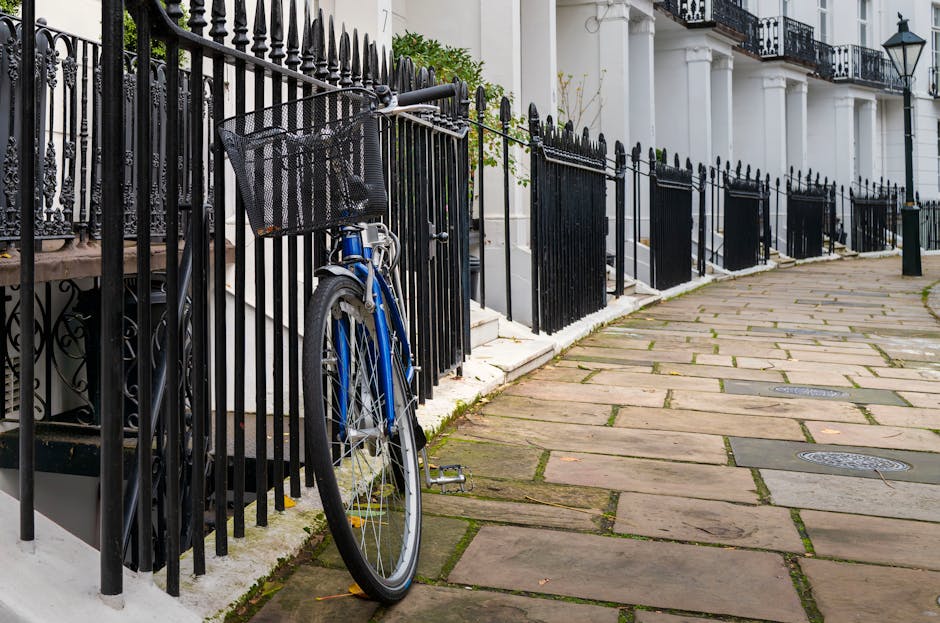 A blue bicycle with a black front basket is locked and propped against a black wrought iron fence along a residential sidewalk in West Kensington. The fence runs parallel to a row of white terraced houses with black metal railings, steps leading up to their entrances, and decorative lampposts mounted on their facades. The sidewalk is paved with large, aged stone slabs, some with visible moss and small leaves scattered across the surface. The scene is lit by natural daylight, highlighting the urban setting typical of Kensington and Chelsea. The bicycle appears to be parked near an area designated for home relocation activities, possibly related to furniture transport or packing and moving logistics, as part of the street's parking restrictions and Van services for removals. The environment suggests a quiet, residential street with an emphasis on handling moving equipment carefully within the designated space, in line with council parking rules.