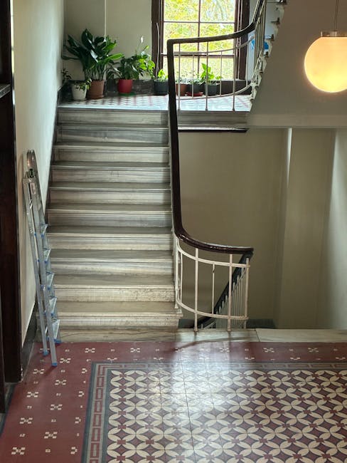 Inside a residential building, a wooden staircase with a dark brown curved handrail leads from the ground floor to the upper level. At the top of the stairs, a small landing features several potted plants placed along the window ledge, allowing natural light to illuminate the area. The flooring on the ground floor consists of decorative patterned tiles in shades of red, white, and gray. To the left of the staircase, a folding aluminium ladder is partially visible, indicating ongoing or recent moving or packing activities. The staircase is narrow, with limited clearance for large furniture, highlighting the challenging access conditions typical for home relocation in tight spaces. The interior wall next to the staircase is painted a neutral light color, and the space is softly lit by a spherical wall light on the right side, creating a calm, neutral environment suitable for professional removal services such as those provided by Man with Van West Kensington.