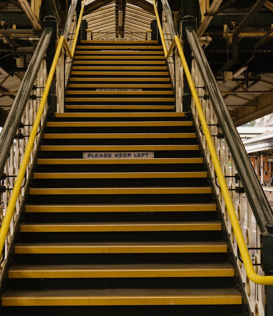 A set of industrial metal staircase with yellow safety handrails leading up within a building, featuring black and yellow non-slip treads, and a white sign on the third step that reads 'PLEASE KEEP LEFT.' The surrounding environment includes structural elements like beams and platforms, suggesting a warehouse or freight area. The lighting is artificial, casting a neutral tone over the scene. This staircase is part of a home relocation or moving process managed by Man with Van West Kensington, facilitating the transport of furniture or appliances in locations with tight stairs or difficult access, aligned with their removals services.