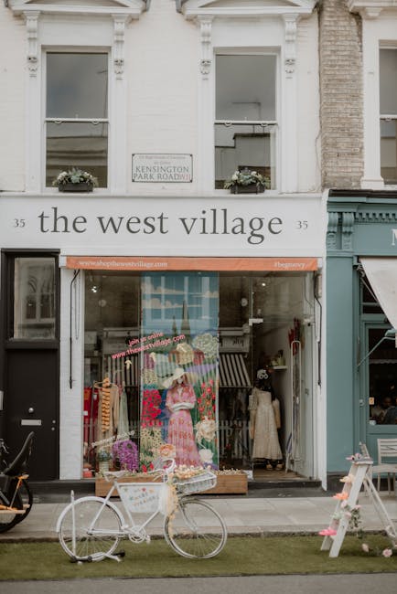 The image shows the façade of a commercial building on Kensington Park Road with two white-framed windows on the upper level, each decorated with flower boxes. The ground floor features a shop named 'the west village' with a display window filled with colorful clothing and mannequins, including a mannequin wearing a pink dress. In front of the shop, on the sidewalk, there is a white bicycle with a basket in front, placed next to a small patch of grass. To the right side of the shop entrance, part of a teal-colored storefront is visible, along with an awning. The scene is illuminated by natural daylight, highlighting the building's details and the street environment. This setting illustrates a typical locale involved in home relocation and moving services, such as those provided by Man with Van West Kensington, especially in the context of packing, furniture transport, and loading processes during house removals.