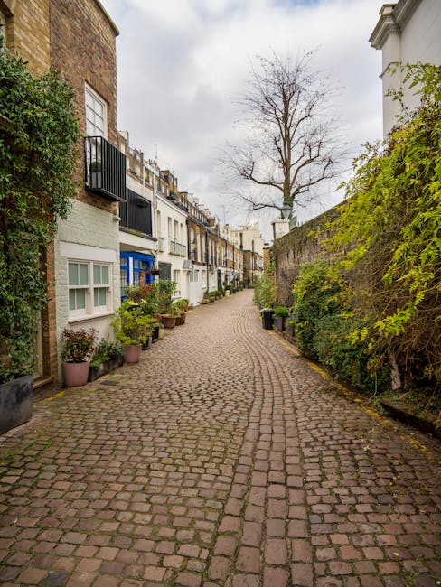 A narrow cobbled street in a residential area with multi-storey houses on the left side, featuring small balconies, potted plants, and a variety of window styles. The street is lined with a mix of greenery, including trees and shrubs, with some leaves showing early signs of autumn. On the right side, a tall stone wall covered partially with green foliage separates the street from adjacent properties. The sky overhead is cloudy, suggesting overcast weather. This scene depicts an urban environment suitable for home relocation or moving logistics, with no moving equipment or vehicles visible, focusing on the residential street context relevant for house removals and furniture transport, as seen in professional removals services like those offered by Man with Van West Kensington.